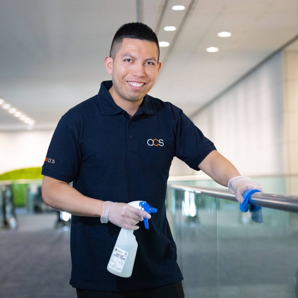 A OCS colleague wearing a shirt and blue gloves is holding a bottle of cleaning solution, ready to clean a surface.