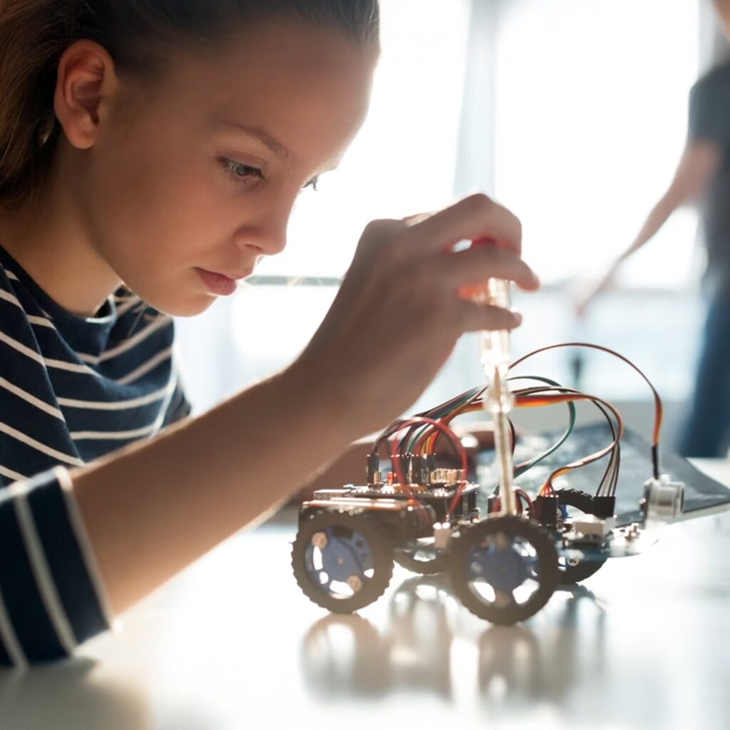 A young person in a striped shirt carefully works on a small wheeled robot, adjusting its wires with focused attention at a well-lit table.