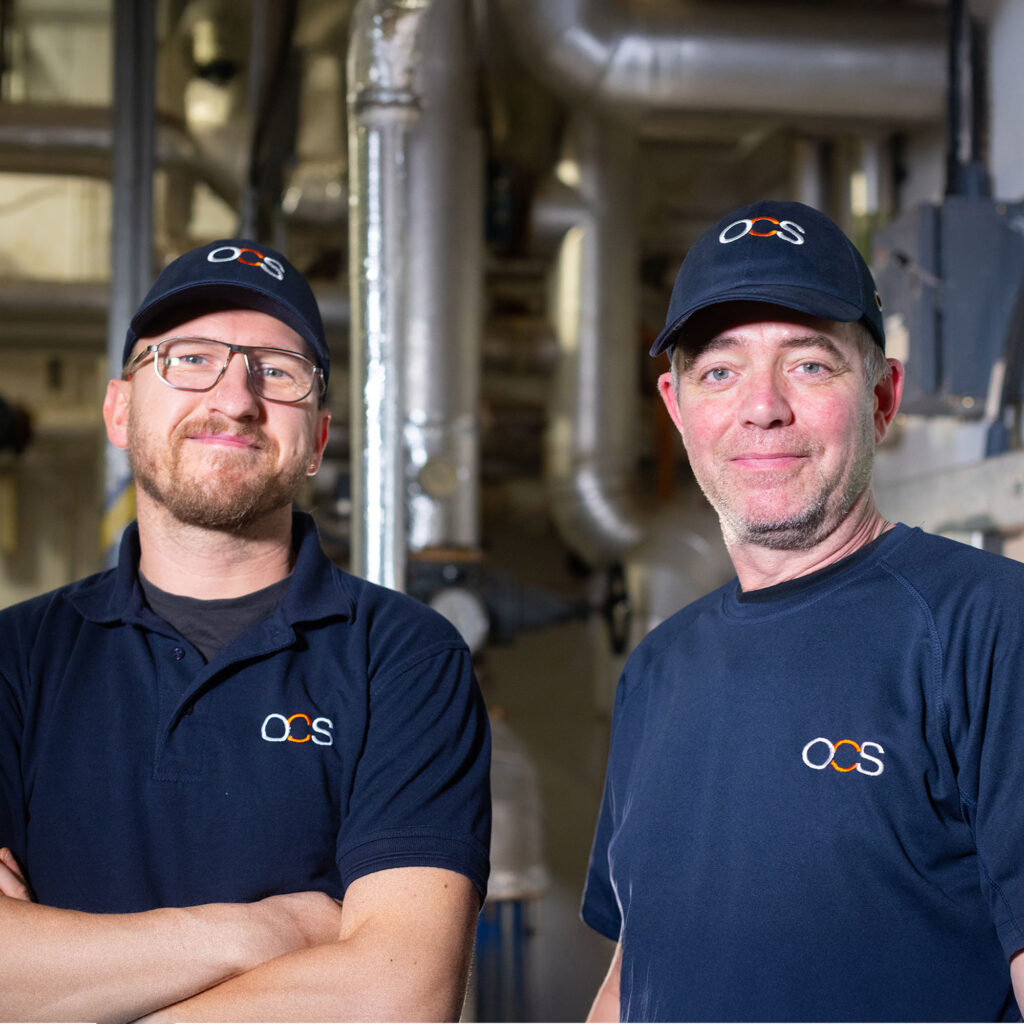 Two men wearing navy blue OCS uniforms and caps stand side by side, smiling, in an industrial setting with visible metal pipes and equipment in the background.