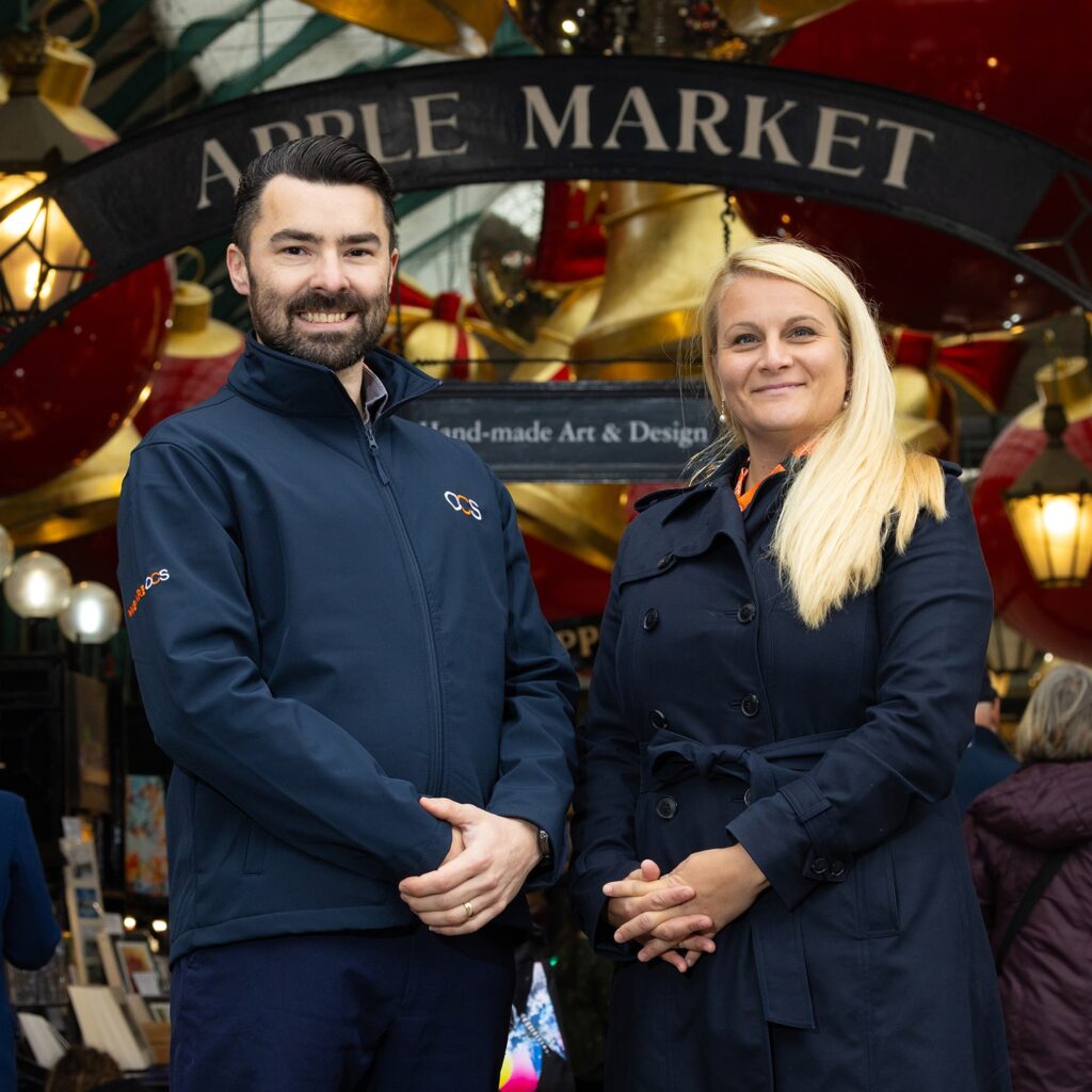 Two people wearing navy jackets stand smiling in front of a decorative Apple Market sign at an indoor market, with warm lights and art stalls visible in the background.