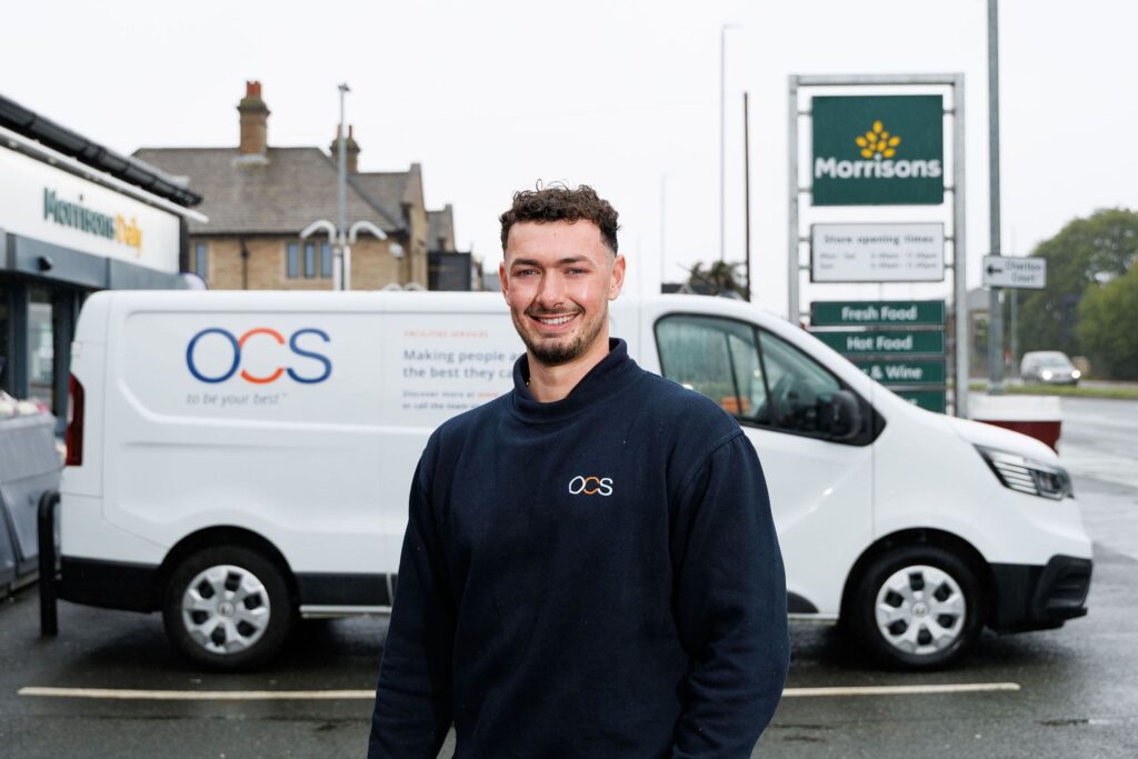 A man in an OCS uniform stands smiling in front of a white OCS van in a Morrisons supermarket parking lot, with a Morrisons sign and store in the background on a cloudy day.
