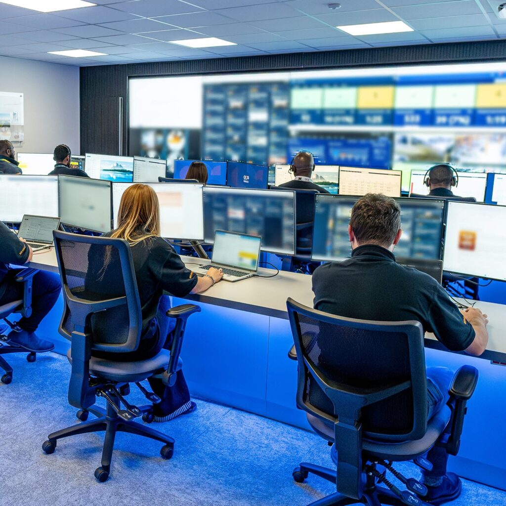 People working at multiple computer stations in a modern control room, facing large wall screens displaying data and images. The room is brightly lit and equipped with ergonomic chairs and headsets.