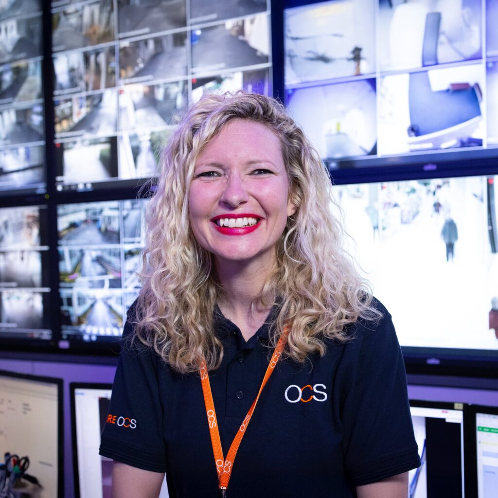 A smiling woman with curly blonde hair, wearing a navy OCS polo shirt and orange lanyard, stands in front of a wall of surveillance monitors displaying various security camera feeds.