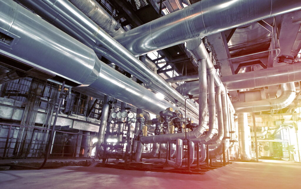 Large industrial facility interior with extensive network of shiny metal pipes, ducts, and machinery. The scene is well-lit, showing gauges and workers wearing hard hats near the center of the image.
