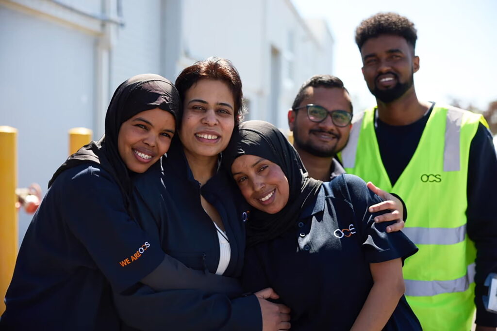 A group of five people, some wearing OCS uniforms, smile and stand close together outdoors on a sunny day. They appear happy and are posing for a group photo in front of a light-colored building.