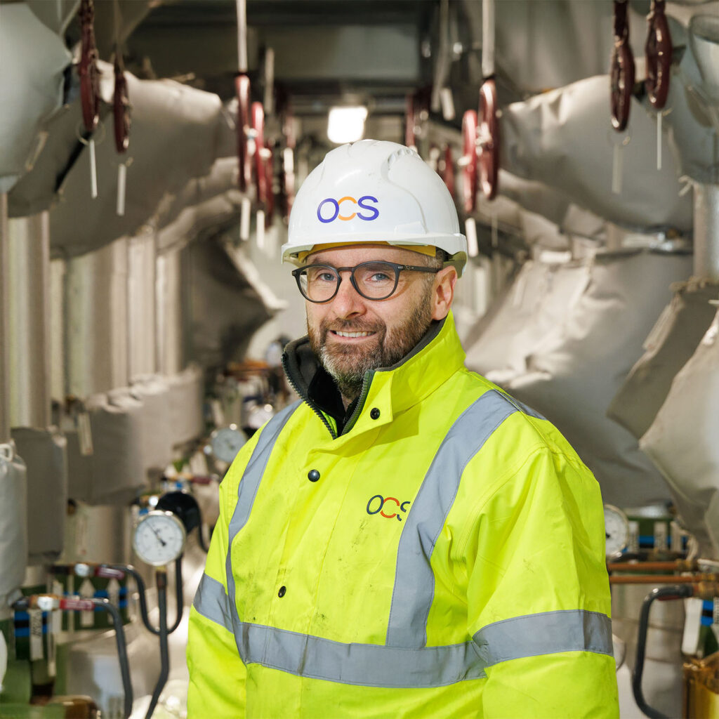 A man in a yellow high-visibility jacket and a white OCS hard hat stands in an industrial setting with gauges and insulated pipes in the background. He is smiling and wearing glasses.
