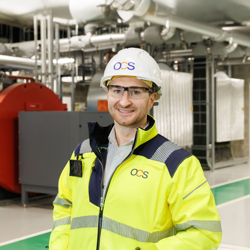 A man wearing a white hard hat, safety glasses, and a yellow high-visibility jacket stands in an industrial facility with pipes and machinery in the background. The hard hat and jacket display the OCS logo.