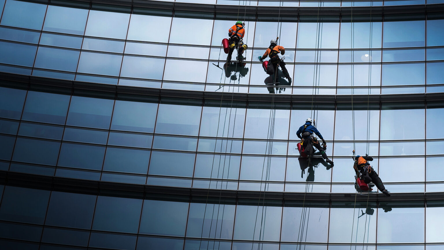 Four workers wearing safety gear clean the windows of a modern, curved glass building while suspended by ropes, with their reflections visible on the glass panels.