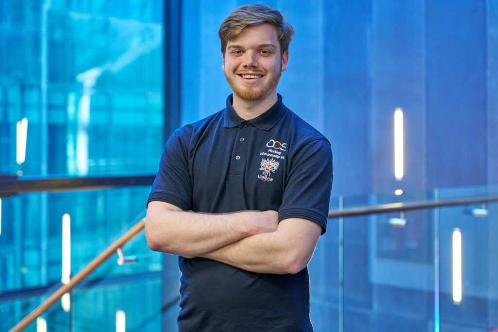A young man with light brown hair and a beard stands smiling with arms crossed, wearing a navy polo shirt with embroidered logos, in a modern building with glass walls and blue lighting.