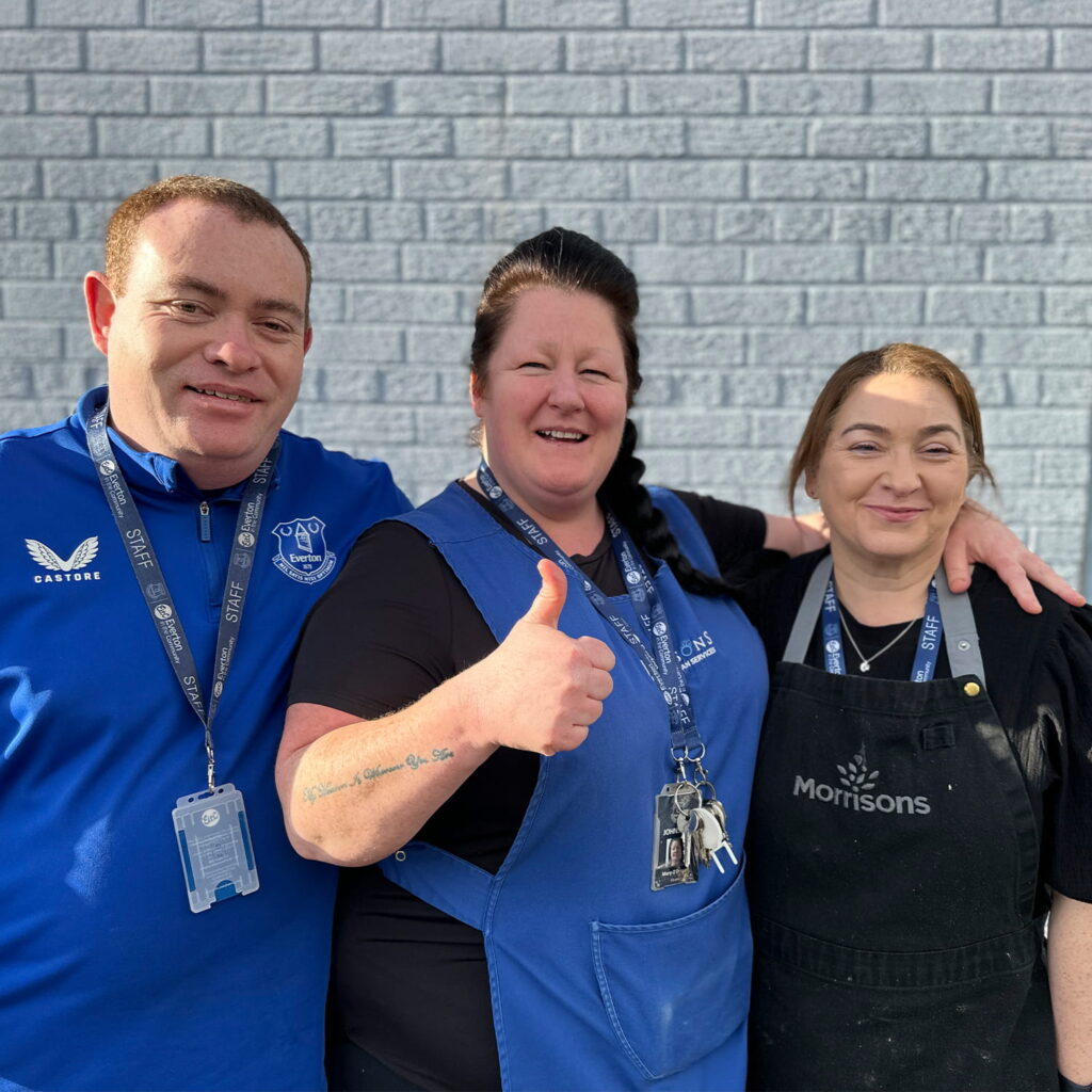 Three smiling people stand together in front of a grey brick wall. The person in the middle gives a thumbs up. Two wear uniforms with lanyards, and the person on the right wears a black Morrisons apron.