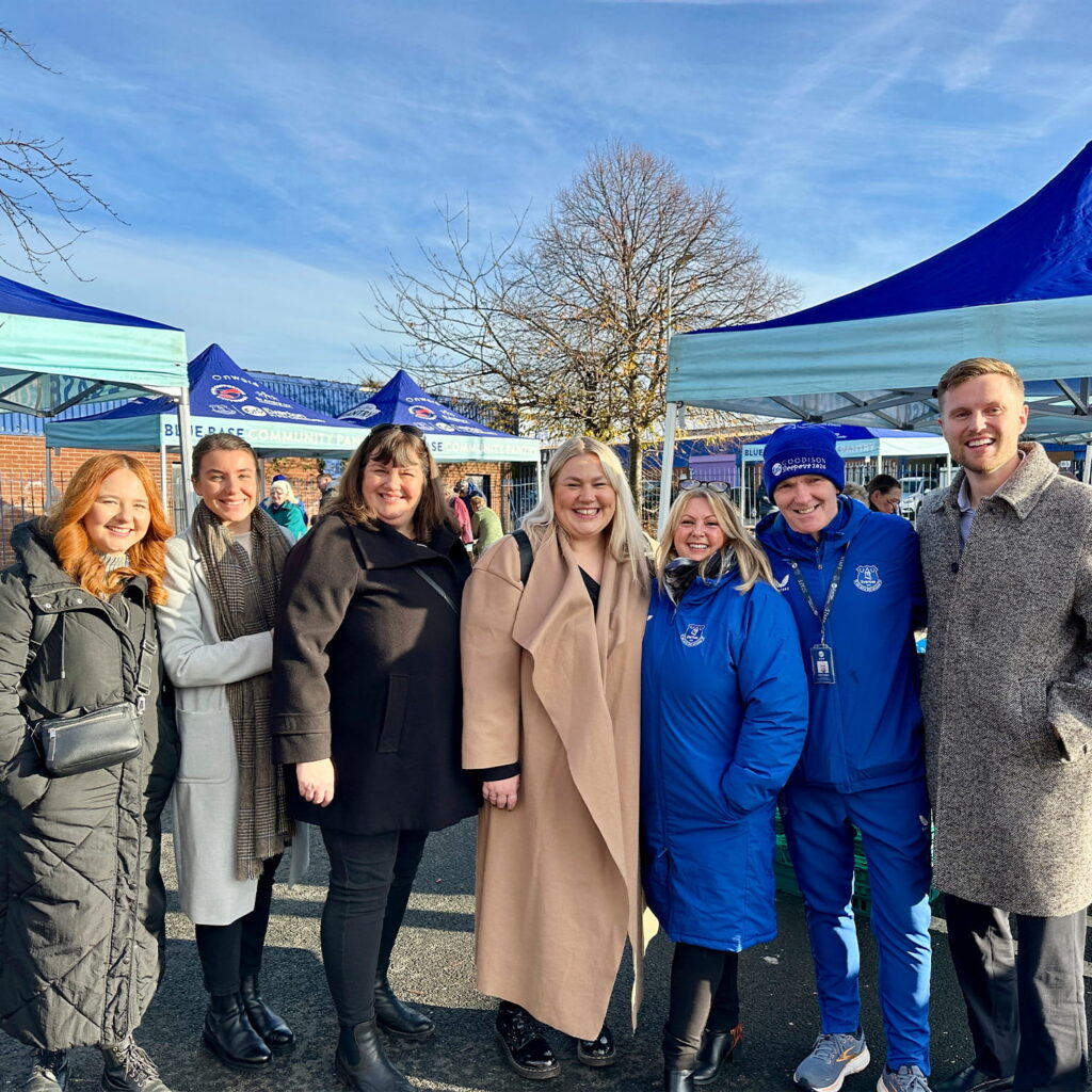A group of seven adults stand smiling together outdoors at an event, with blue and white tents and leafless trees in the background. Some wear coats, and a few are dressed in blue sportswear. The weather is sunny.