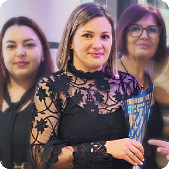 Three women stand together indoors. The woman in front is holding a decorative trophy and wearing a black, lace-patterned dress. The two women behind her are also dressed in black. All are facing the camera and smiling slightly.
