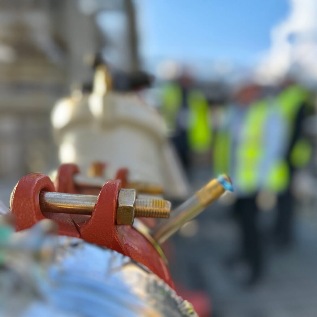 Close-up of a metal pipe with bolts and red clamps in focus, while several people wearing yellow safety vests and hard hats stand blurred in the background at what appears to be a construction site.