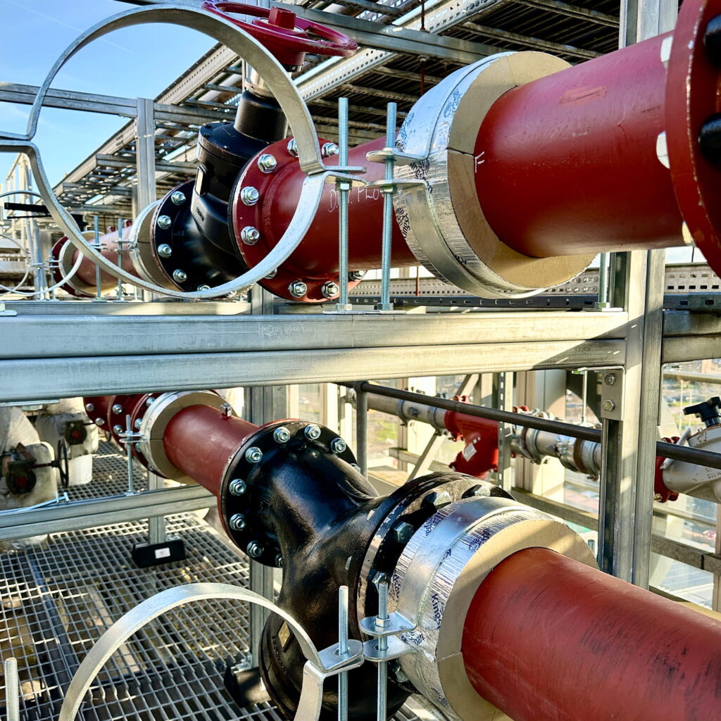 Red and black industrial pipes secured with metal brackets run parallel on a metal grate platform, surrounded by a metal framework and outdoor industrial equipment.