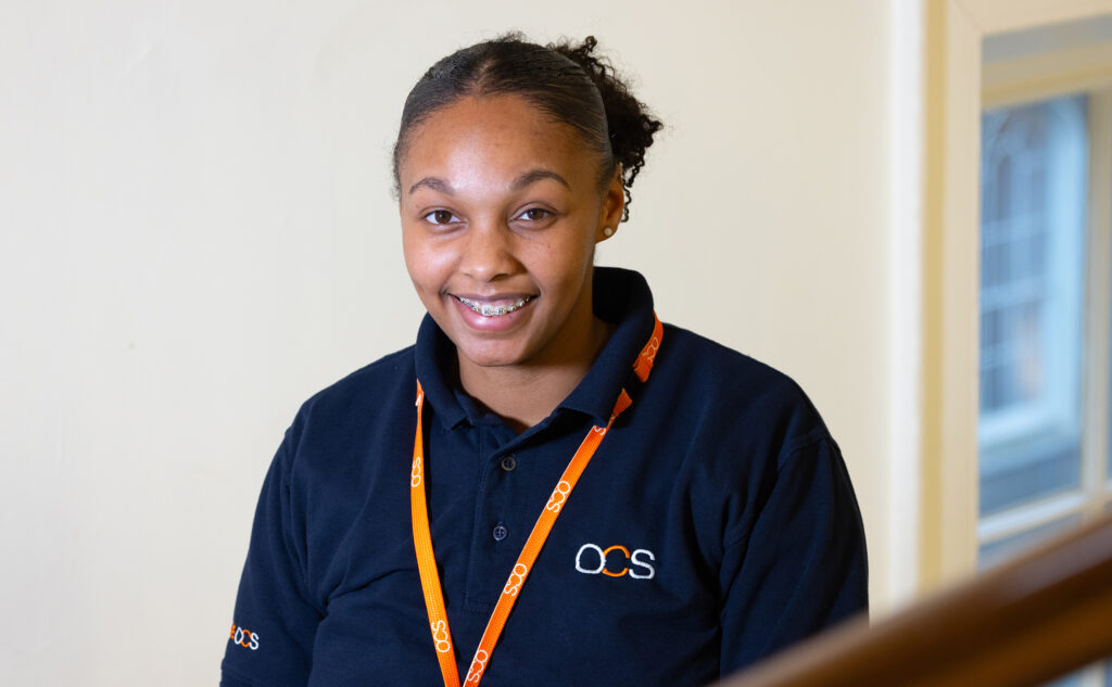 A woman wearing a navy blue OCS uniform and orange lanyard smiles, standing indoors near a staircase with a light-colored wall and window in the background.