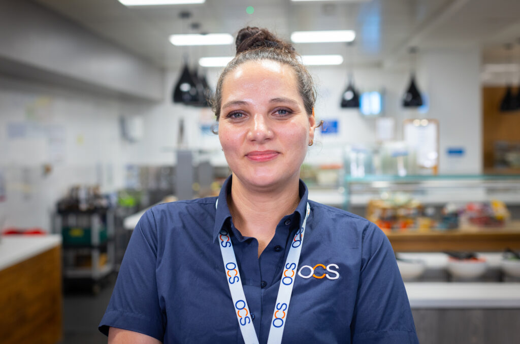 A woman with brown hair in a bun, wearing a navy blue OCS uniform and lanyard, stands in a bright, modern cafeteria with food displays and kitchen equipment in the background.