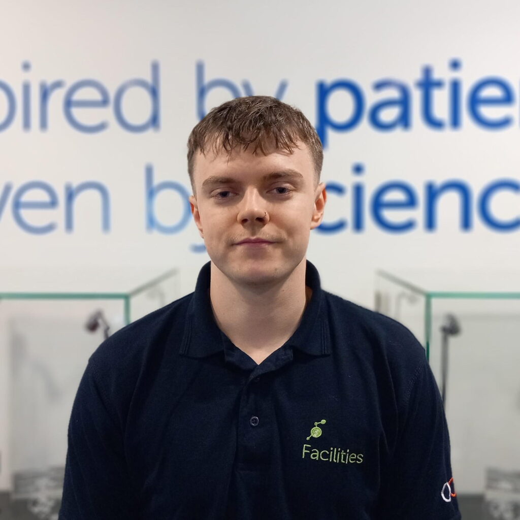 A young man with short brown hair, wearing a navy blue Facilities polo shirt, stands indoors in front of a blurred wall featuring large text with the words patients and science.
