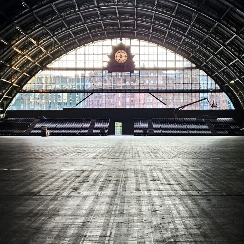 Wide, empty indoor hall with a large arched glass window, a clock at the center above the window, and rows of seating along the sides beneath exposed steel beams. Sunlight streams in from outside.