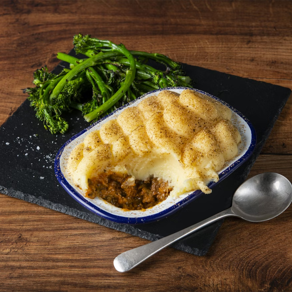 A serving of shepherd’s pie with a golden mashed potato topping in a small dish, part of it scooped out, is placed on a slate tray with steamed broccolini and a spoon on a wooden table.