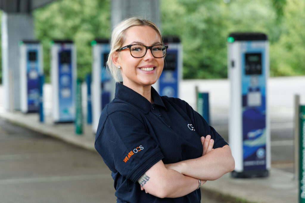 A smiling woman with glasses and a ponytail stands with arms crossed at an electric vehicle charging station, wearing a navy polo shirt with an orange WIRELOGIC logo on the sleeve. Blurry green trees are in the background.