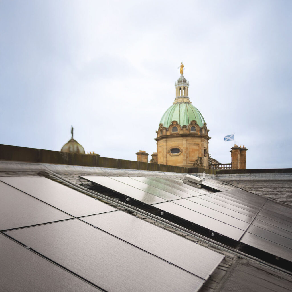 Solar panels on a rooftop in the foreground, with a historic building featuring a green dome and a statue under a cloudy sky in the background. A small flag is visible near the dome.