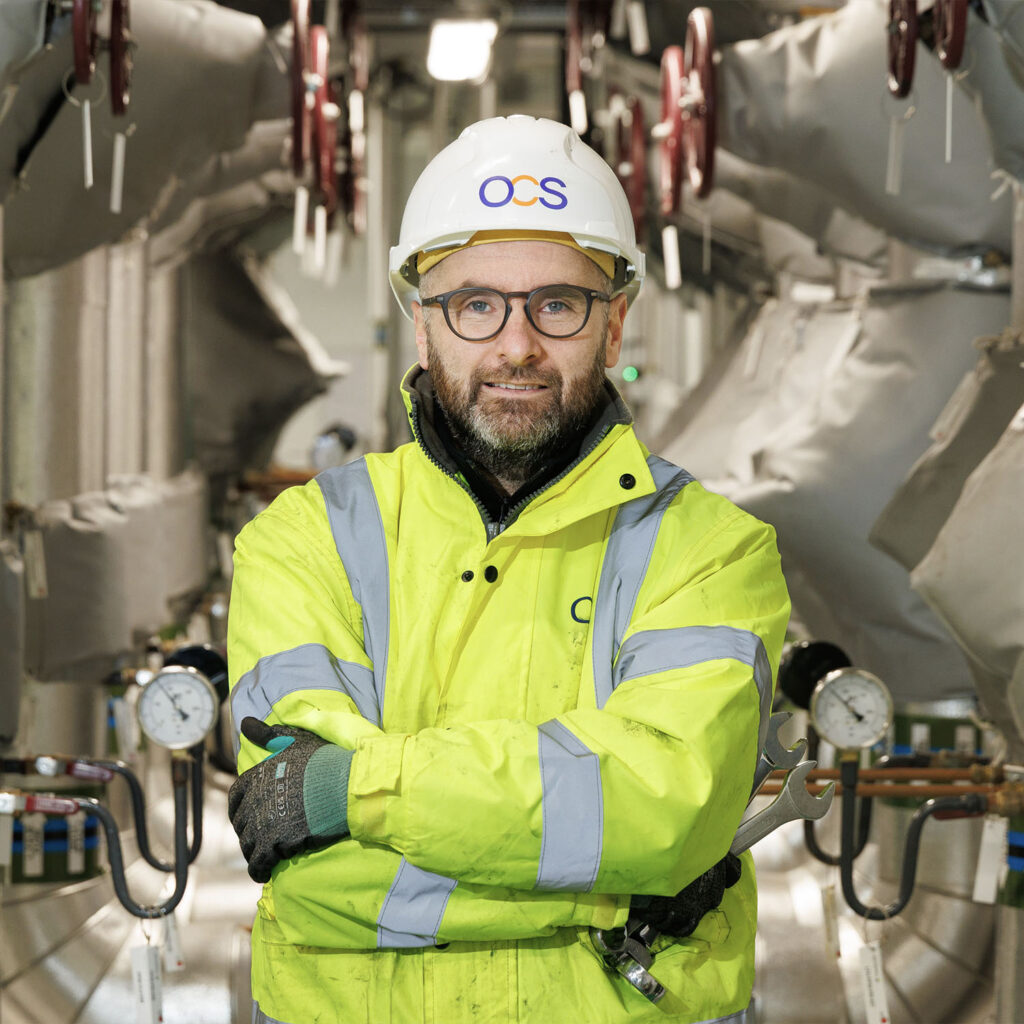 A man wearing a safety helmet and high-visibility jacket stands with arms crossed in an industrial setting, surrounded by pipes, gauges, and equipment.