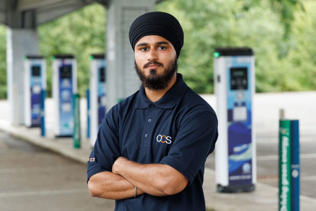 A man wearing a black turban and a navy blue ODS polo shirt stands with folded arms in front of several electric vehicle charging stations outdoors.
