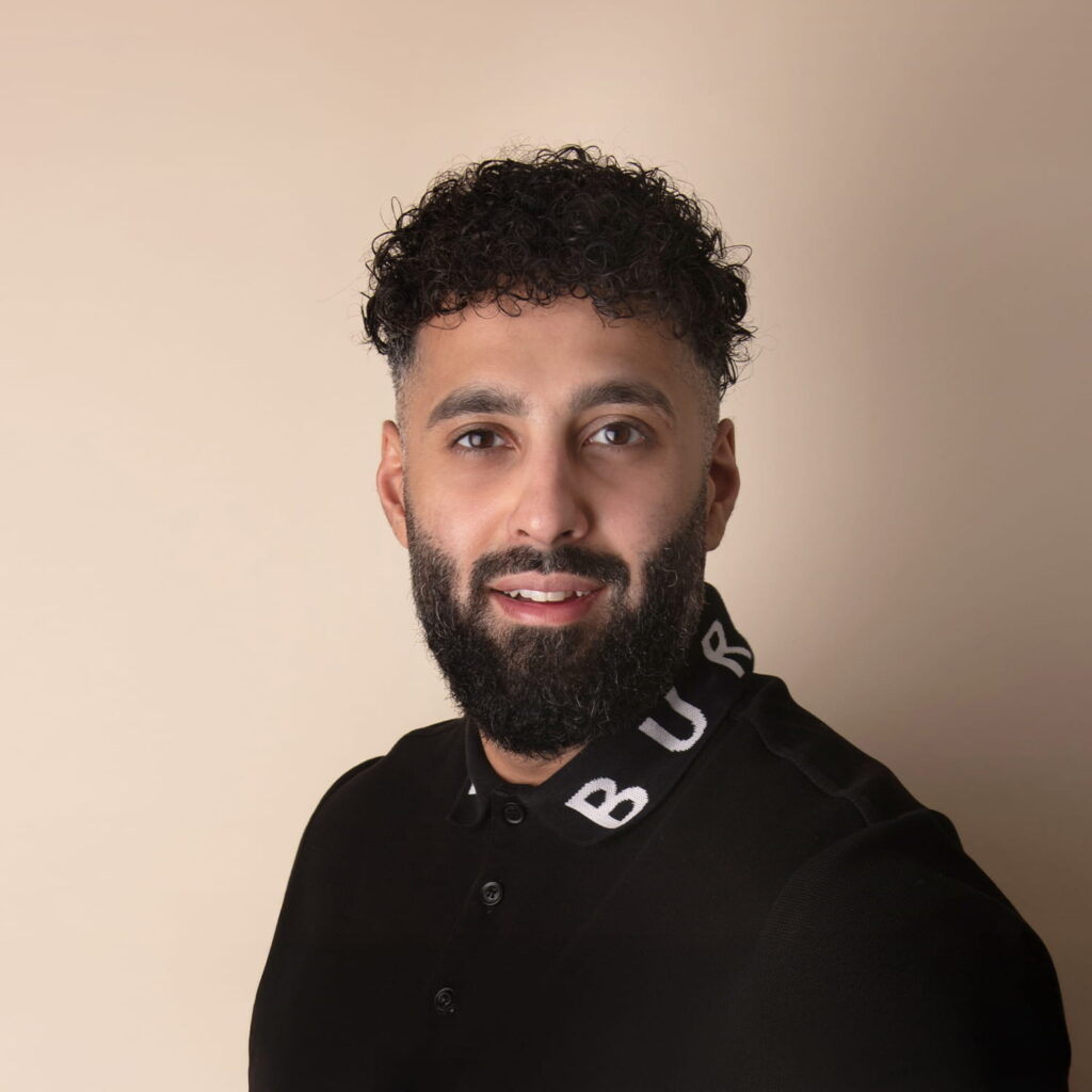 A man with short curly black hair and a beard smiles at the camera. He is wearing a black collared shirt with the letters BUR visible on the collar, standing against a plain beige background.
