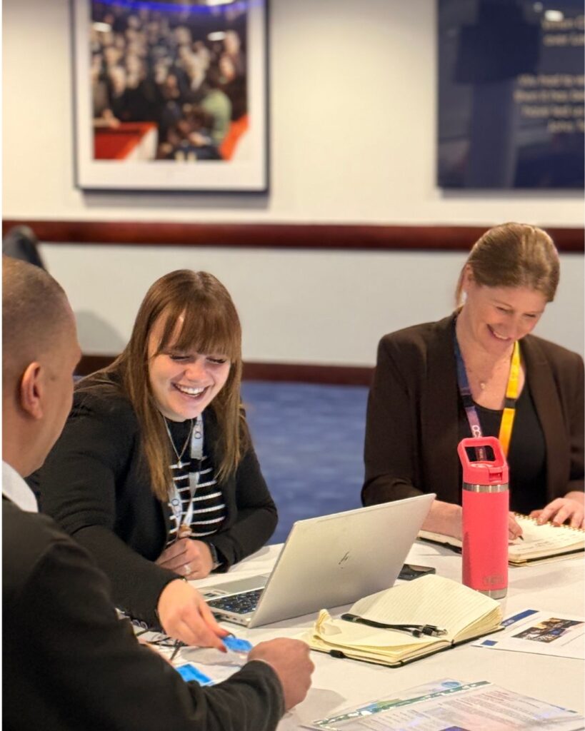 Three people sit at a table working and smiling, with laptops, notebooks, and a pink water bottle in front of them. Two women and one man are engaged in conversation in a bright, professional setting.
