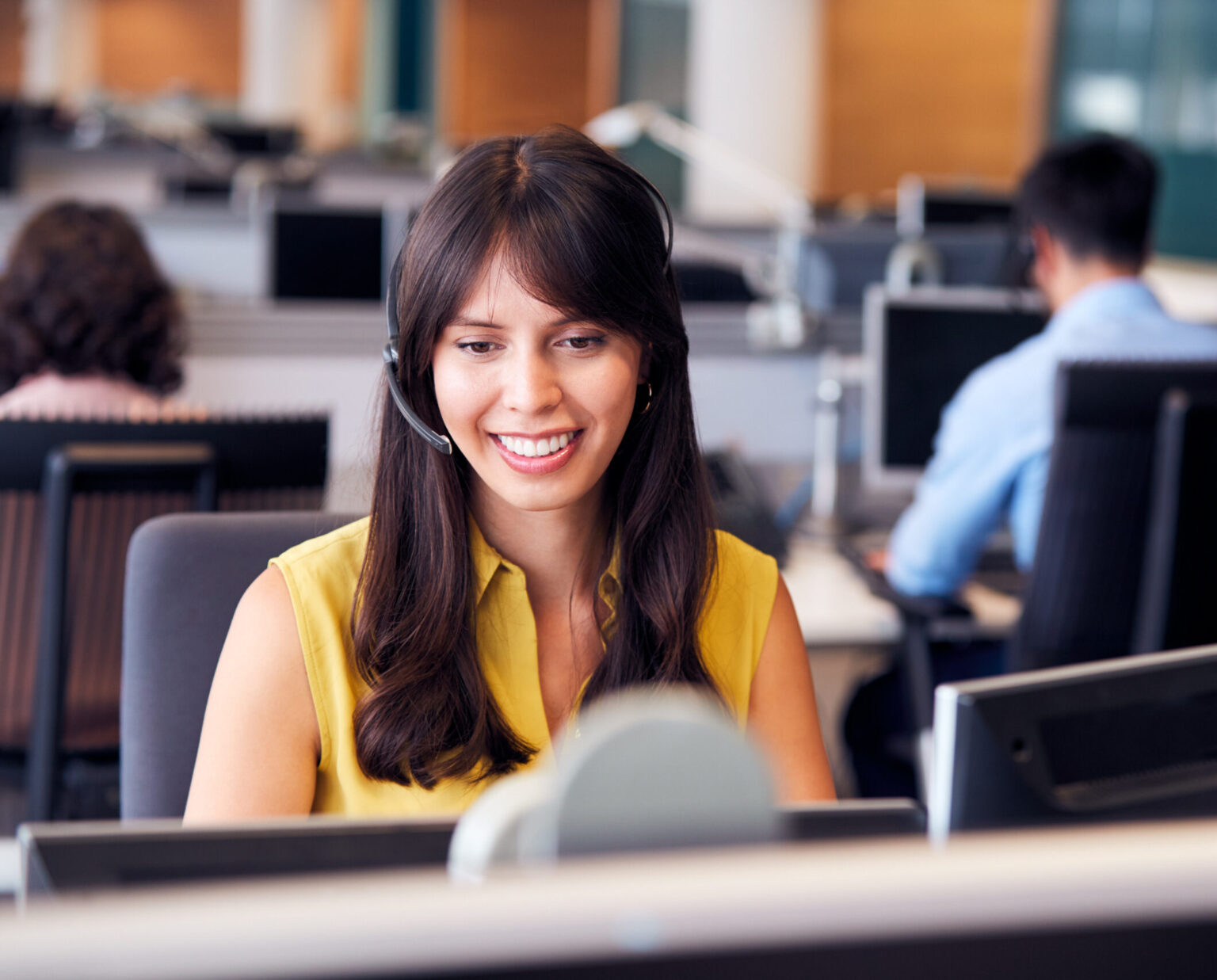 Woman wearing a headset and a yellow sleeveless blouse smiles whilst working at a computer in a modern office with colleagues in the background.