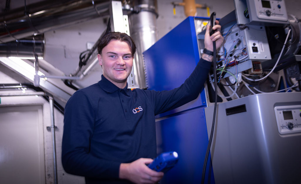 A technician in a navy blue shirt smiles whilst working on electrical equipment in an industrial setting, holding a tool and cables near open control panels.