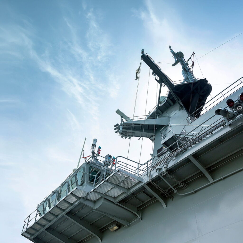 The image shows the upper deck and command tower of a naval aircraft carrier against a blue sky with wispy clouds. Aerials, railings, and equipment are visible on the ship’s structure.