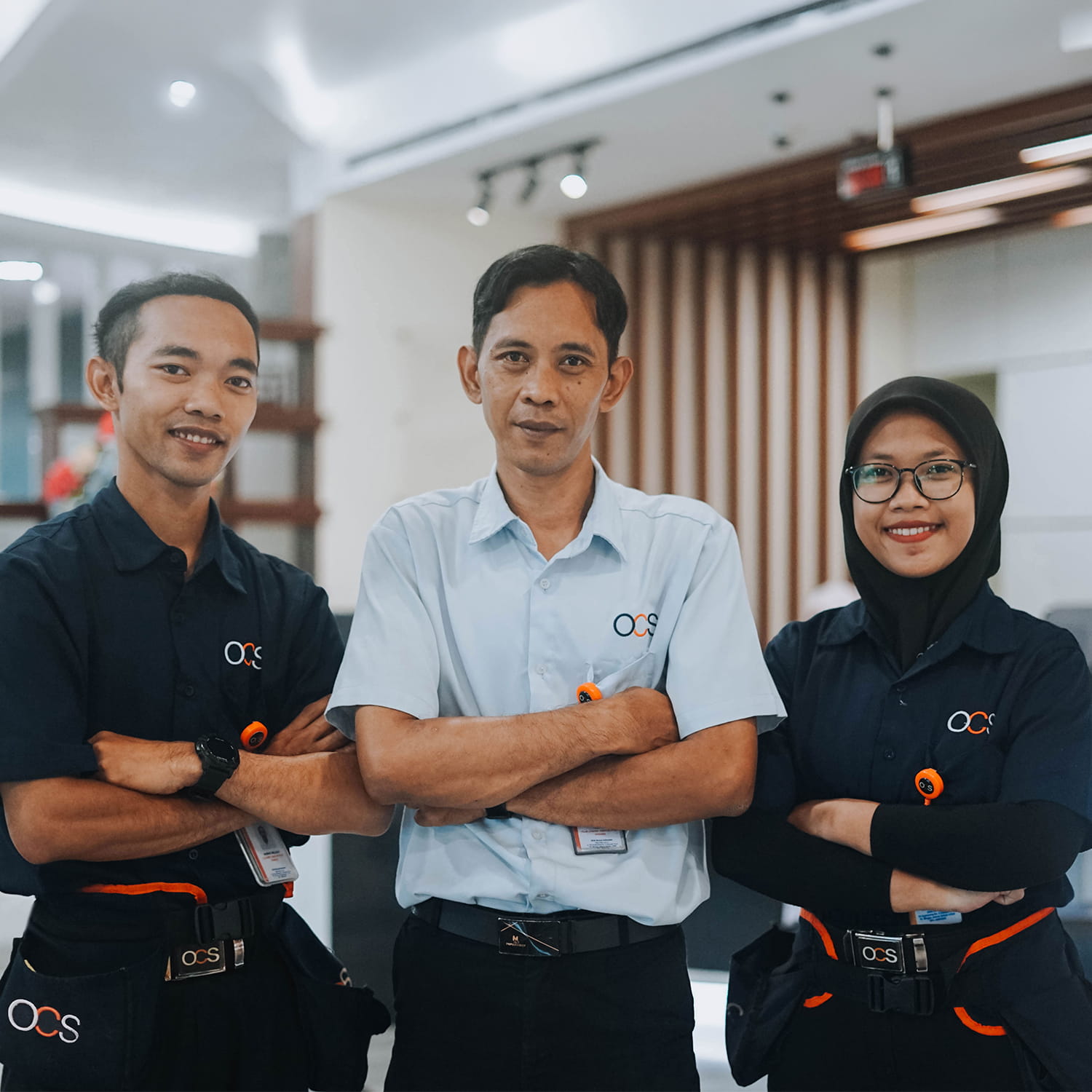 Three employees in uniform stand side by side indoors, smiling confidently with arms crossed. Two men and one woman, all wearing OCS-branded shirts and ID badges, pose in a modern office setting.