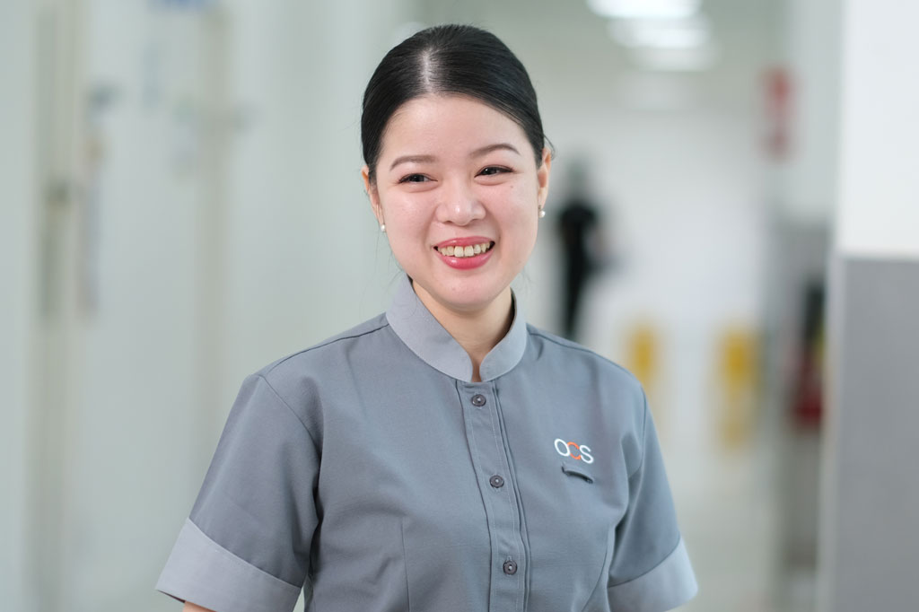 A woman in a gray uniform with a company logo smiles while standing in a bright hallway. The background is slightly blurred, showing a corridor with a few indistinct figures.