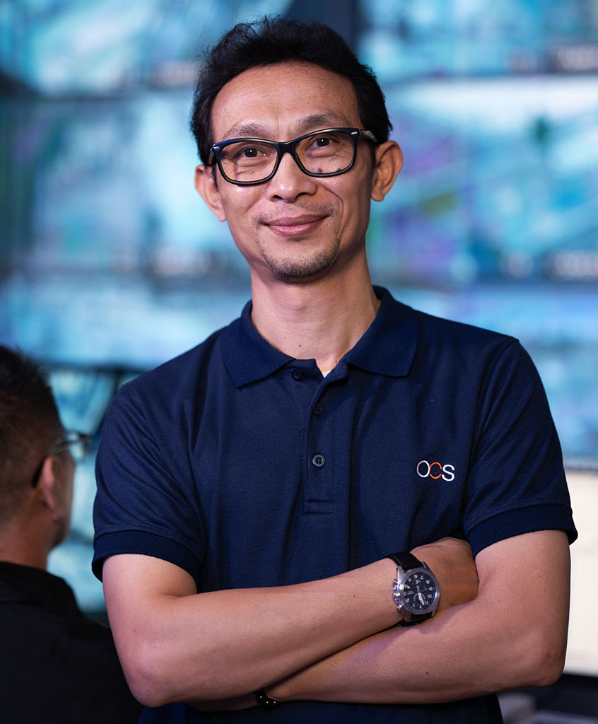 A man wearing glasses and a navy polo shirt with an OCS logo stands with arms crossed, smiling. He is in front of monitors with blue screens, giving a professional, tech-savvy impression.