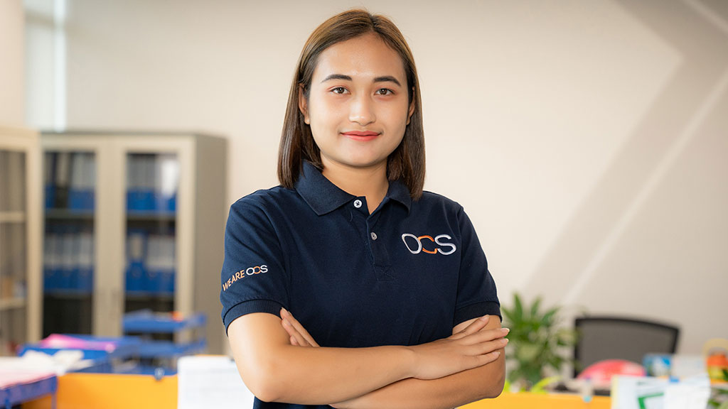 A woman with straight brown hair stands in an office, wearing a navy blue polo shirt with the OCS logo, arms crossed, and smiling confidently at the camera. Shelves with folders are visible in the background.