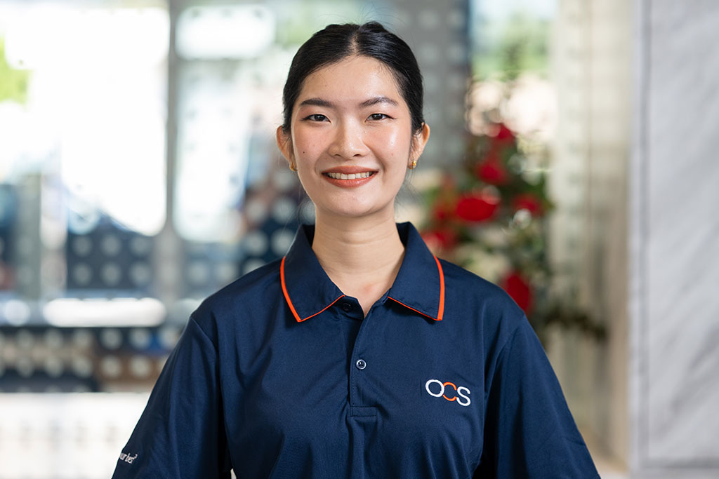 A woman wearing a navy blue OCS polo shirt smiles at the camera, standing indoors with a blurred background featuring some red flowers.