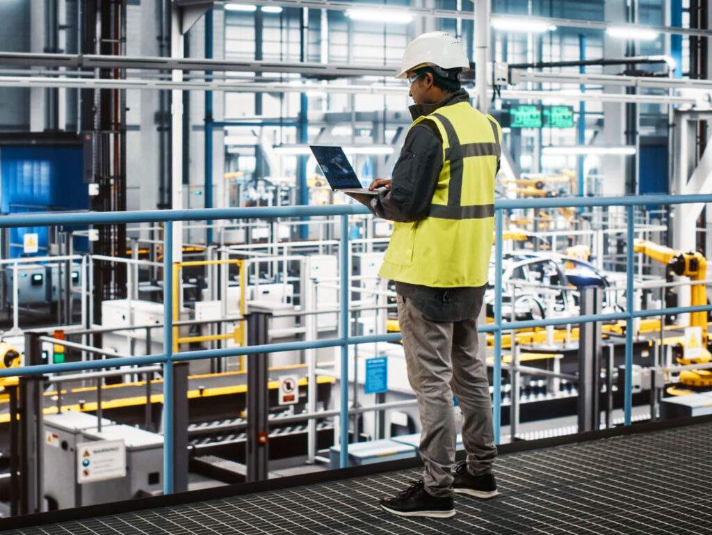 A person in a hard hat and yellow safety vest stands on a platform in a factory, using a laptop while overlooking industrial machinery and equipment.