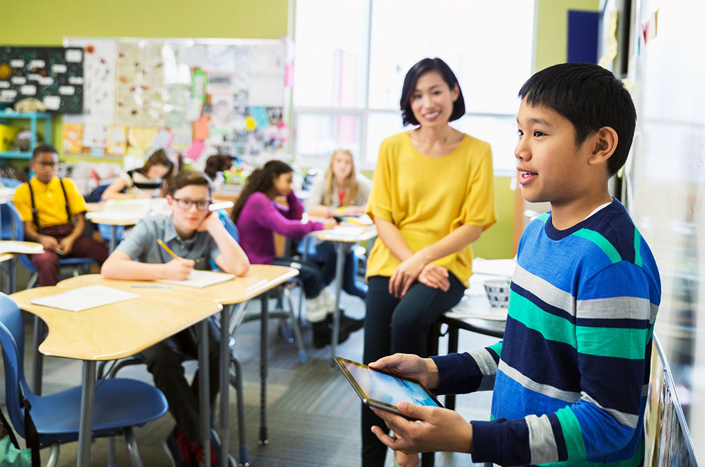 A boy stands at the front of a classroom holding a tablet, speaking to his classmates. Other students sit at desks, and a teacher in a yellow top smiles in the background. The classroom is bright and colorful.