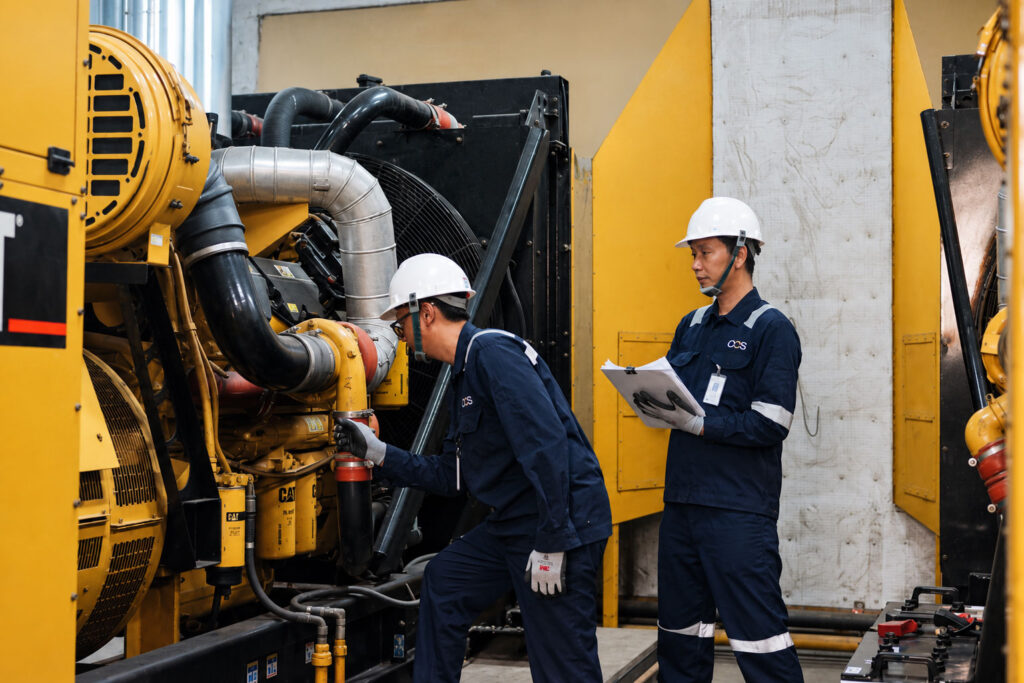 Two workers in blue uniforms and white helmets inspect and take notes on a large yellow industrial machine in a facility. One operates the equipment, while the other observes and writes on a clipboard.
