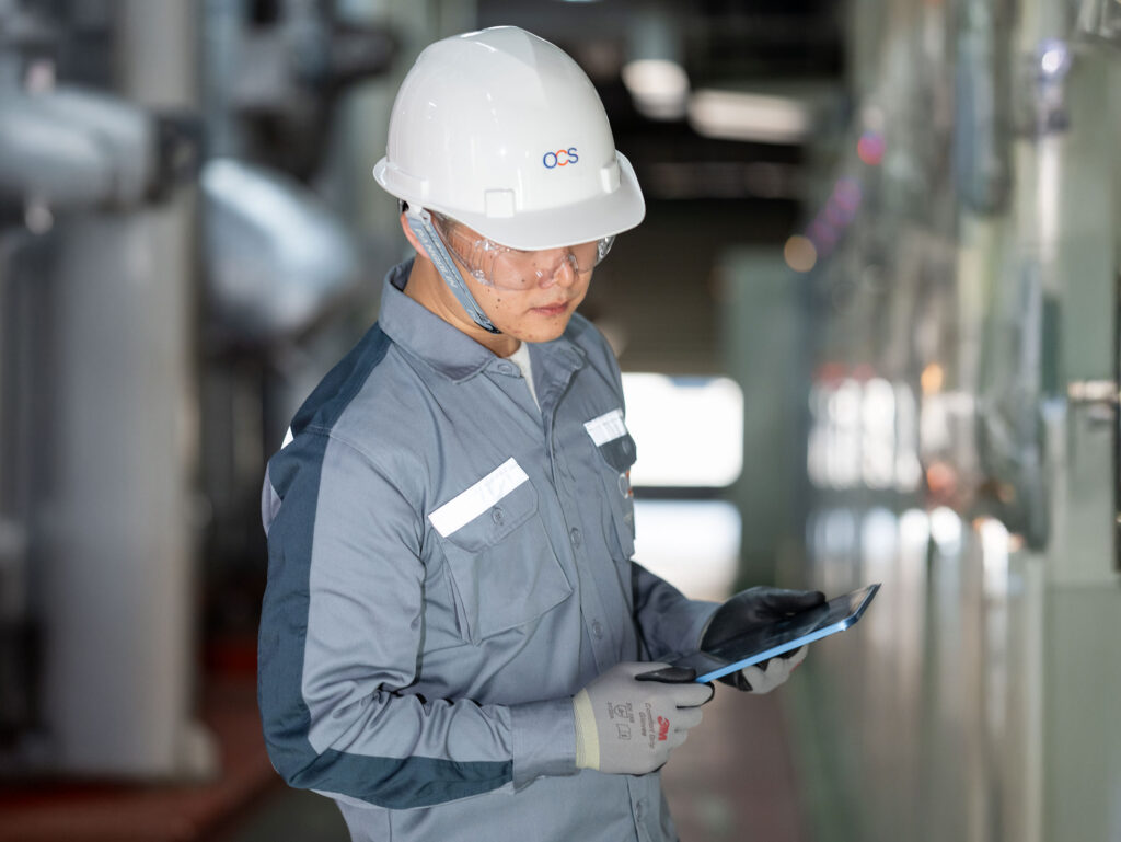 A worker in safety gear, including a helmet, gloves, and goggles, uses a tablet while inspecting equipment in an industrial facility.
