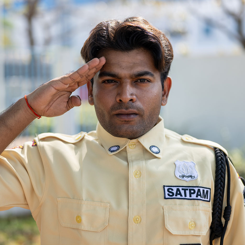 A security guard in a yellow uniform labeled SATPAM stands outdoors, giving a salute with his right hand. The background is blurred.