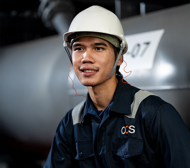 A person wearing a white safety helmet and a dark blue work uniform with OCS on the chest stands in an industrial setting.