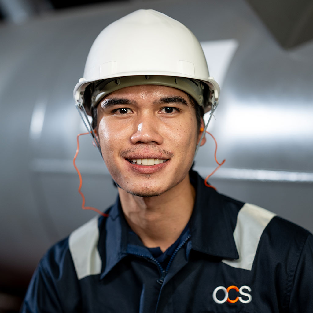 A man wearing a white hard hat and a dark blue uniform with an OCS logo smiles at the camera, standing in an industrial setting.