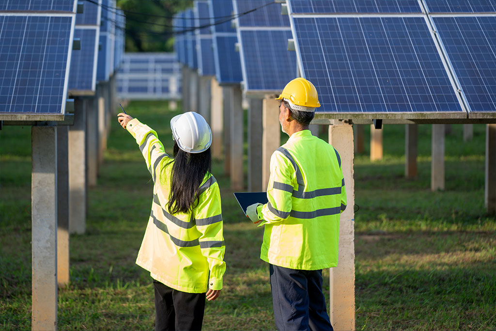 Two workers in safety gear inspect solar panels outdoors; one points towards the panels while the other holds a clipboard. They are surrounded by rows of solar panels on a grassy field.