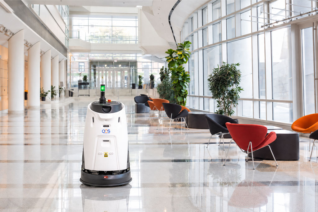 A white cleaning robot moves through a modern, spacious lobby with large windows, colorful chairs, and green plants.