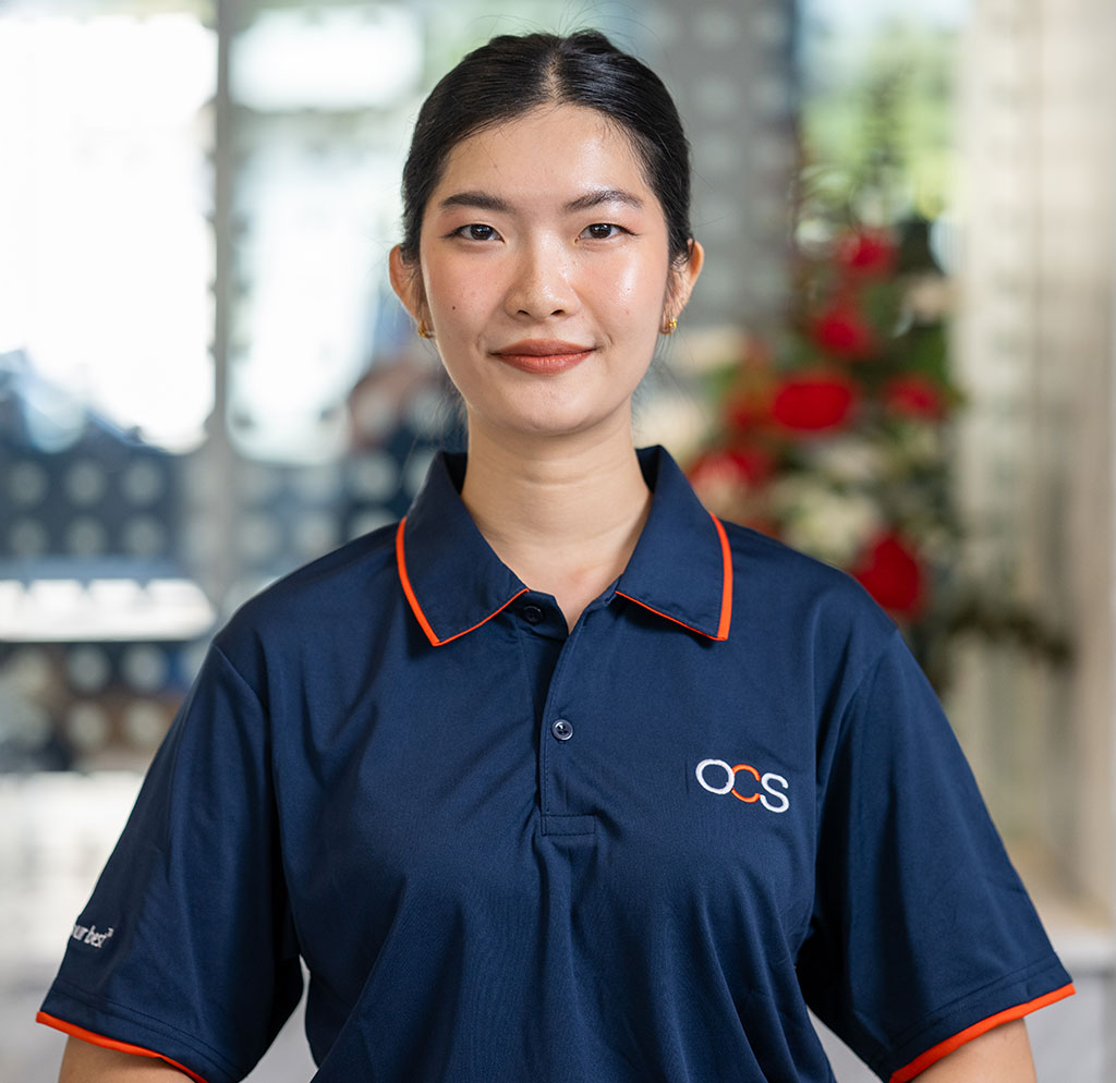 A woman wearing a navy blue OCS polo shirt stands indoors, smiling at the camera. There are blurred flowers and a glass wall in the background.