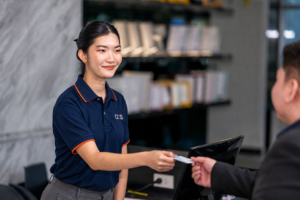 A receptionist in a navy blue shirt smiles as she receives an ID card from a person, standing at a desk with office shelves in the background.