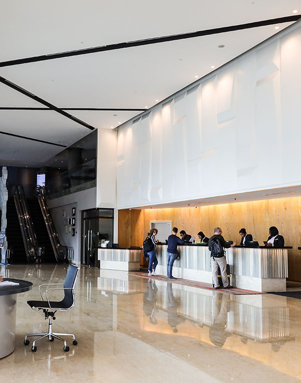 Modern hotel lobby with people standing at a long reception desk, staff assisting guests, shiny marble floor, and escalators in the background. An empty office chair sits near the foreground.