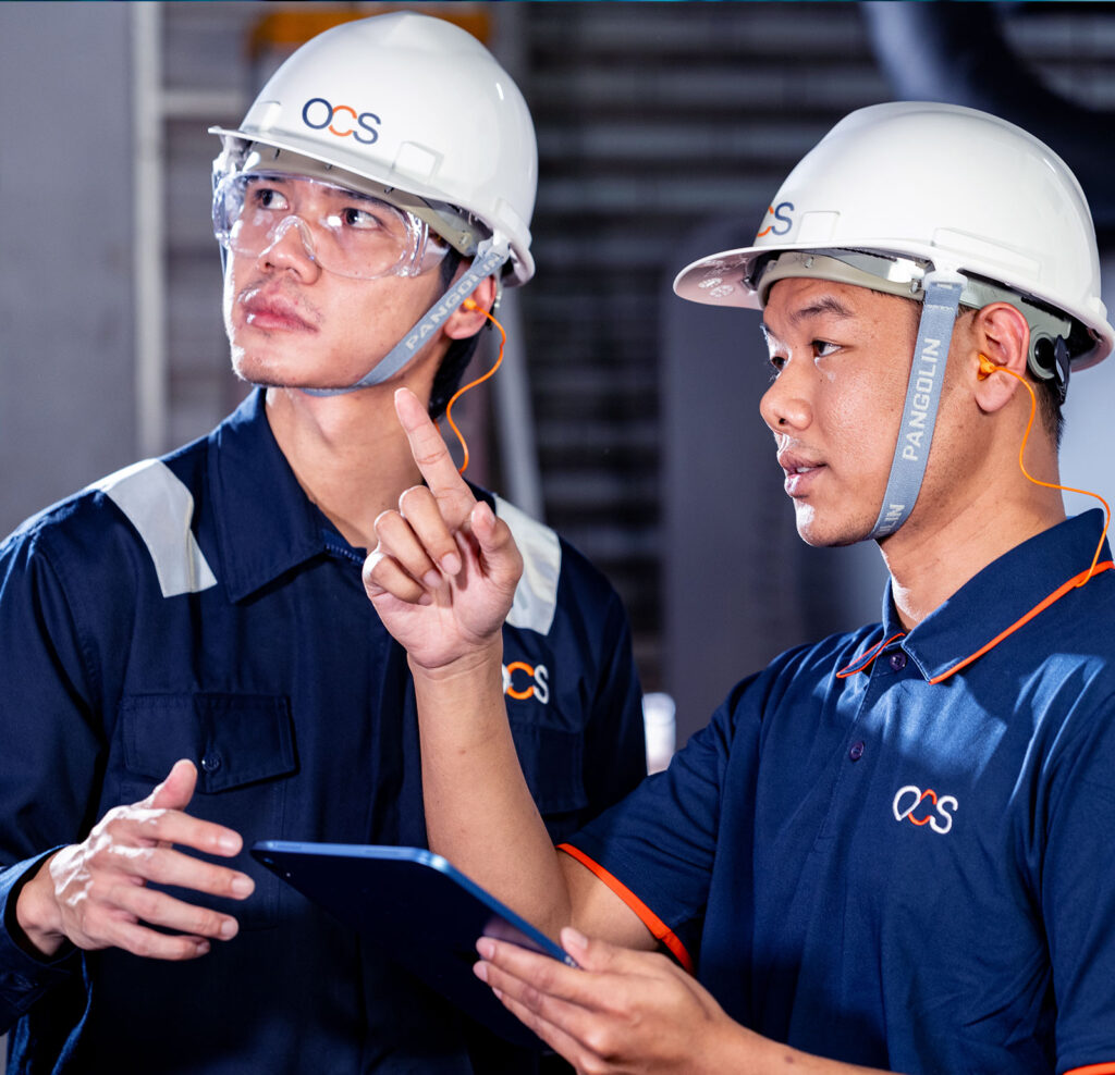 Two workers in blue uniforms and white hard hats discuss something while one points and the other listens. Both wear safety glasses and ear protection, and one holds a tablet.
